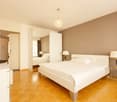Spacious bedroom with white walls, hardwood floors, a large bed, bedside tables with lamps, and a crystal chandelier. There is a closet and a doorway leading to another room.