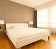 Spacious bedroom with white walls, large windows, hardwood floor, white bed frame, two bedside tables with lamps, and curtains covering the window