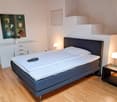 The image shows a bedroom with a double bed, a dresser, and a side table. The room has white walls, hardwood floors, and is well-lit with lamps. There is a framed artwork on the wall above the dresser.
