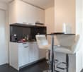 Compact kitchen with white cabinets, black granite countertop, built-in dishwasher, and bar seating area with two white leather stools
