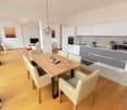 The image shows an open-plan living area with a dining table, chairs, and a kitchen. The dining table has a wooden top and metal legs. The chairs are beige in color. The kitchen has white cabinets and drawers, with a gray countertop. There is a dishwasher