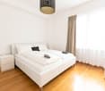 Spacious bedroom with white walls, hardwood flooring, a large bed with white bedding, a nightstand, and a patterned pendant light fixture. The room also features a curtained window, suggesting the presence of a balcony or terrace.