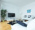 Spacious living room with white walls, hardwood floors, large windows, white sofa, glass coffee table, TV stand, and a potted plant.