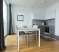 The image shows a modern, open-plan kitchen and dining area. The kitchen features grey cabinets and appliances, including a dishwasher. The dining table is a simple white rectangular table with four chairs. The room has large windows providing plenty of n