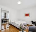 Spacious bedroom with hardwood floors, white walls, large window, bed, nightstand, artwork, and a vase of flowers on a table.