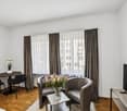 Spacious living room with large windows, hardwood floor, dining table, armchairs, and a vase of flowers on a side table