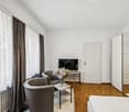 Spacious bedroom with large windows, hardwood floor, gray armchairs, coffee table, and a TV stand. The room has a modern and minimalist design.