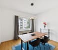 wooden table and chairs, blue rug, white walls, window with view, radiator, shelf with flower