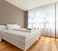Spacious bedroom with white bed frame, white bedding, and large windows with sheer curtains providing natural light. The room has hardwood flooring and a neutral color scheme.