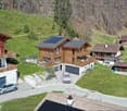 Wooden chalet style houses, 3 garages, solar panels on roofs, driveway, cars parked in front, landscaped area with flowers and plants, surrounding trees and mountains.