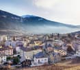 Panoramic view of a mountainous city with colorful buildings, surrounded by snow-capped peaks and hazy mountains in the background