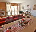 Cozy living room with wooden walls, large windows, red sofa, wooden shelves and cabinets, decorative items like candles and pine cones, and a plaid tablecloth on the coffee table.