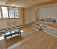 Open space, wooden floor, natural light from window, cozy seating area with pillows, coffee table, radiator on wall, wooden handrail