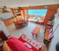 Open living room with wooden dining table, living room couch, and balcony with view. Wooden cabinets, tiled flooring, decorative items, and lamps are present. Kitchen area with stove and microwave. Dining table set with chairs. Balcony has table, chairs,