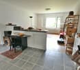 The image shows an open-plan living area with a kitchen, dining, and living room. The kitchen features a granite countertop, a dishwasher, and various kitchen appliances. The living room has a sofa and shelving units. The floor is tiled, and there is a wi