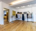 a room with white walls, wooden floor, clothes hanging on racks, clothes hanging on a wall, a fan, a cabinet, and pictures on the wall