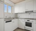 Bright and modern kitchen with white cabinets, granite countertops, tiled backsplash, and stainless steel appliances including a dishwasher. Large windows provide natural light.