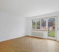 Spacious room with large windows overlooking a garden, hardwood parquet flooring, and a sliding glass door leading to a balcony or terrace.
