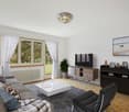 Spacious living room with large windows, white curtains, hardwood floors, gray sofa, coffee table, TV stand, and shelving unit. The room has a modern and minimalist design with a balcony or terrace visible through the windows.