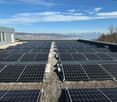 The image shows a large array of solar panels installed on the roof of a building. The solar panels are arranged in rows and cover a significant portion of the roof. The background shows a scenic view of a city and mountains, indicating that the building
