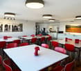 The image shows a large open dining or breakroom area with multiple tables and chairs. The tables have a white surface and the chairs are red in color. The room has modern lighting fixtures and artwork on the walls, including some landscape photos. There