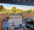 covered terrace, large couch, two chairs, coffee table, two hanging lamps, garden view, golf course in the distance