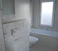 A clean and minimalist bathroom with white tiles, a bathtub, a toilet, a sink, a mirror, and a window. Modern fixtures.