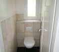 The image shows a bathroom with a white toilet, tiled walls, and a small window. The floor is covered with dark tiles.
