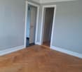 Empty room with gray walls, hardwood herringbone flooring, and two doorways visible.