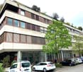 Office building, beige exterior, glass windows, 4 stories, green plant life on rooftop, street view, cars parked