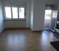 The image shows an open-plan living room and kitchen area. The room has large windows that provide natural light and access to a balcony or terrace. The floor is made of hardwood or laminate flooring. The kitchen area is equipped with modern appliances.