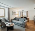 Large windows, gray sofa, armchair, wooden coffee table, rug, wooden floor, lamp, picture frame, dining table, chairs