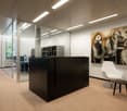 Modern and minimalist office space with glass walls, black furniture, and a large abstract painting on the wall. The space has recessed lighting and a comfortable-looking white chair.