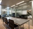 Large open office space with glass walls, modern furniture, and artwork on the walls. The room has a long conference table with leather chairs, and there are additional workstations visible through the glass walls.