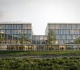 Modern office building with glass facade, multiple floors, large windows, entrance with glass doors, people walking around, trees, and grass in front.