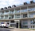 Multi-story apartment building with modern, minimalist design. The building has several balconies and a parking area in front. The exterior features a combination of glass, concrete, and metal elements.
