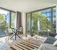 Spacious living room with large windows overlooking a balcony and a view of the city. The room features a dining table, chairs, coffee table, and a sofa. The decor is modern and minimalist, with a striped rug on the hardwood floor.