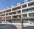 Multi-story apartment building with modern, minimalist design. The building features balconies on each floor, with a mix of glass and wood accents. There is a covered entrance area at the ground level.