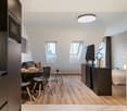 Open-plan studio apartment with kitchen, dining area, and living space. Hardwood flooring, large windows, modern appliances including a dishwasher, and a TV stand.
