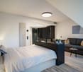 Spacious bedroom with a king-size bed, a dark wood TV stand, a sofa, and a modern lighting fixture. The room has a minimalist and contemporary design with neutral colors and wood flooring.