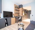 Modern living room with a large TV, wooden shelving unit, desk area, and kitchen visible in the background. The room has a minimalist and contemporary design with a gray and wood color scheme.
