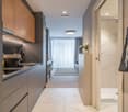 Modern kitchen with wood and gray cabinets, stainless steel appliances, and a marble-like countertop. The kitchen leads to a hallway with a striped rug and curtains.