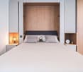 Spacious bedroom with wooden paneled walls, a large bed with gray bedding, and a built-in wardrobe with lighting. There is also a small shelf unit next to the bed.