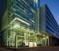 Large modern glass and steel office building with a prominent entrance and parking area in front. The building has multiple floors and a sleek, contemporary design with a lot of natural light coming through the glass facade.