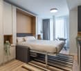 Spacious bedroom with a large bed, built-in wardrobes, and a striped rug on the hardwood floor. The room has large windows with curtains, providing natural light and a potential balcony or terrace access.