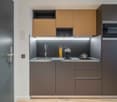Compact kitchen with modern, minimalist design. Features wood-tone upper cabinets, gray lower cabinets, stainless steel appliances including a dishwasher, and a sleek countertop with a sink and cooking utensils.