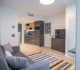 Open-concept studio apartment with hardwood floors, gray sofa, kitchen with stainless steel appliances including a dishwasher, and a TV stand.