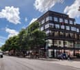 A multi-story commercial building with a modern, black and glass facade. The building has several shops and businesses on the ground floor, with residential or office spaces on the upper floors. There are people walking on the sidewalk in front of the bui