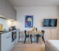 Modern studio apartment with open kitchen, dining area, and living space. The kitchen features gray cabinets, a stove, and a dishwasher. There is a small dining table with two chairs, and a large TV mounted on the wall. The room has a minimalist, contempo