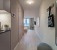 Compact studio apartment with kitchenette, hardwood floors, large windows, and a bedroom area at the end of the hallway. The apartment features a dishwasher and a tote bag hanging on the wall.